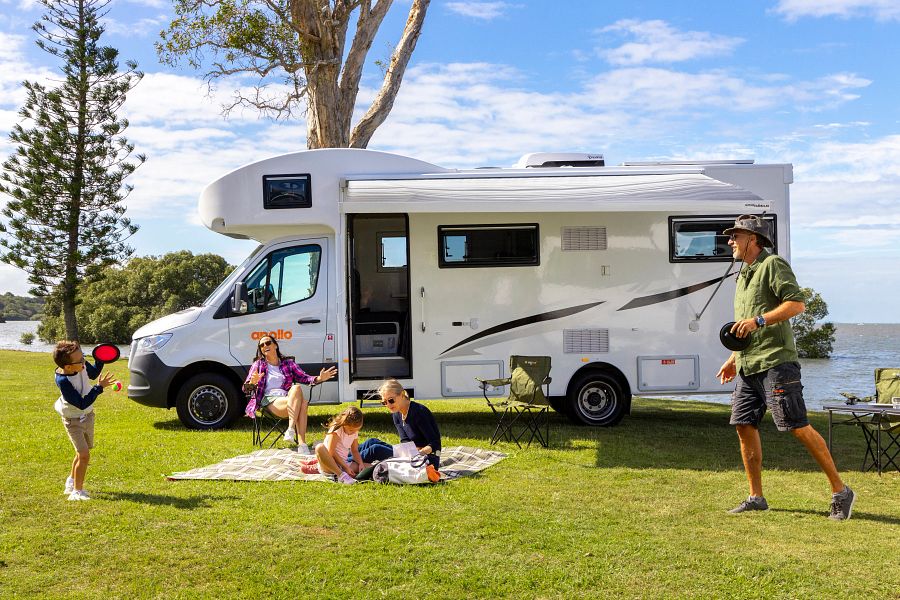 Gezin speelt buiten voor de Apollo Euro Deluxe camper, vader speelt kleefbal met zoon, moeder zit met dochter op picknickkleed