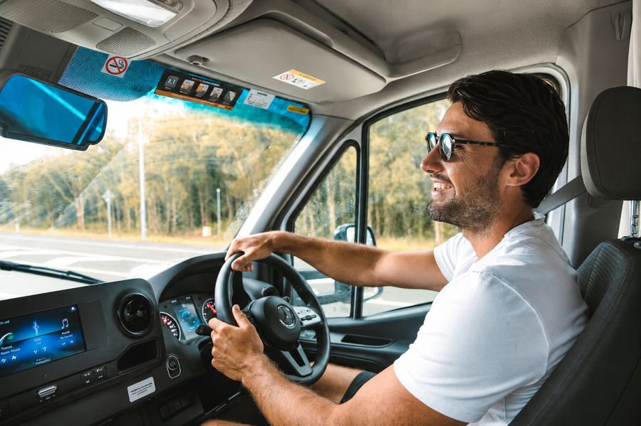 Man rijdt in Mighty Big Six camper, lacht naar camera, zonnebril op