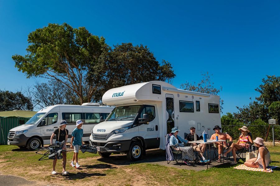 Gezin met Maui Beach camper zit buiten op de camping, twee tieners komen aanlopen met nog meer stoelen