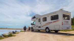 Cruisin' Discovery camper staat geparkeerd op het zand aan een meer, stel zit op campingstoelen buiten