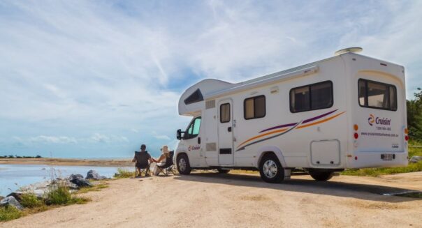 Cruisin' Discovery camper staat geparkeerd op het zand aan een meer, stel zit op campingstoelen buiten