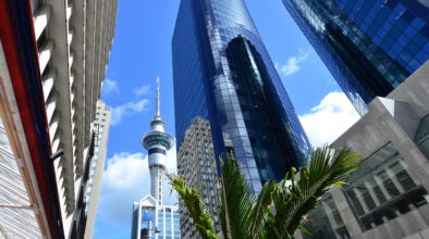 Zicht op de Skytower in Auckland van onderaf gefotograafd, met groene palm op de voorgrond en wolkenkrabbers eromheen