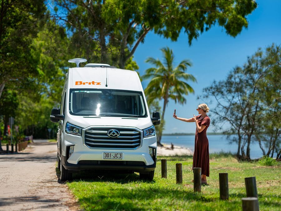 Vrouw maakt selfie naast de Britz Trailblazer in Queensland die bij het strand geparkeerd staat tussen palmbomen
