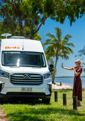 Vrouw maakt selfie naast de Britz Trailblazer in Queensland die bij het strand geparkeerd staat tussen palmbomen