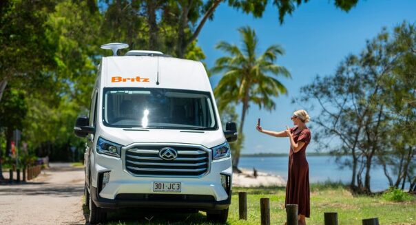 Vrouw maakt selfie naast de Britz Trailblazer in Queensland die bij het strand geparkeerd staat tussen palmbomen