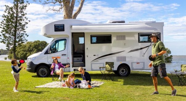 Gezin speelt buiten voor de Apollo Euro Deluxe camper, vader speelt kleefbal met zoon, moeder zit met dochter op picknickkleed