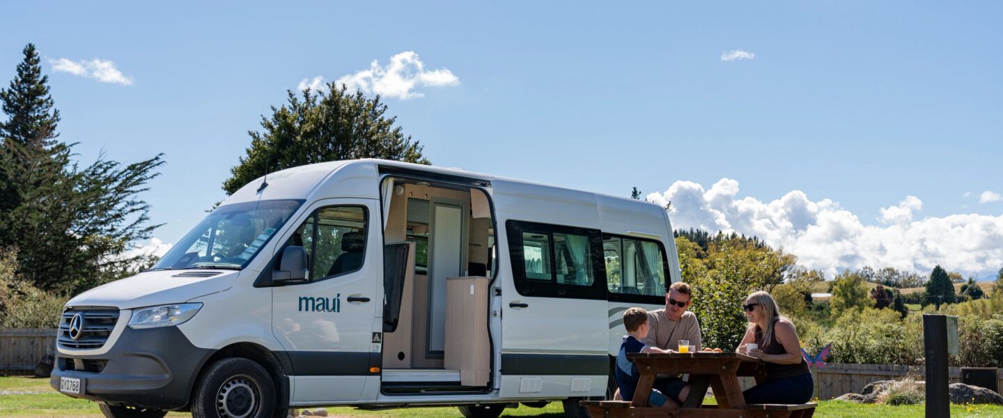 Vader, moeder en kind zitten aan picknicktafel voor de Maui Ultima Plus camper bij Lake Taupo in Nieuw-Zeeland