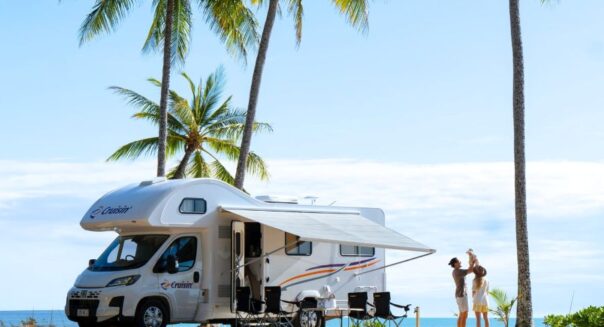 Vader tilt jong kind op naast Cruisin' 6 Berth Deluxe camper die tussen palmbomen aan het strand geparkeerd staat