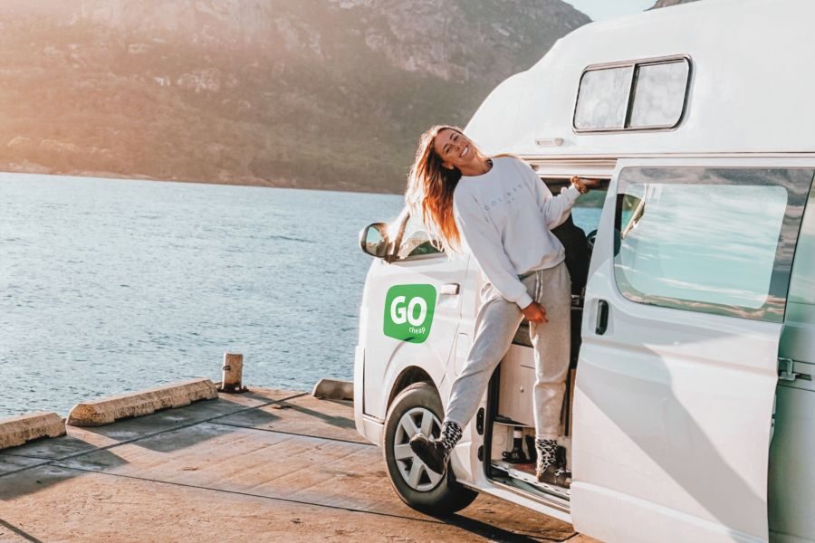 Een vrouw hangt uit de deur van de Go Cheap 4 Berth Hitop die geparkeerd is aan het water