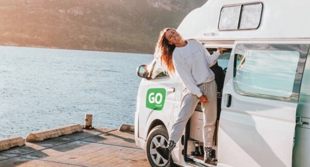 Een vrouw hangt uit de deur van de Go Cheap 4 Berth Hitop die geparkeerd is aan het water