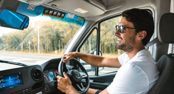 Man rijdt in Mighty Big Six camper, lacht naar camera, zonnebril op