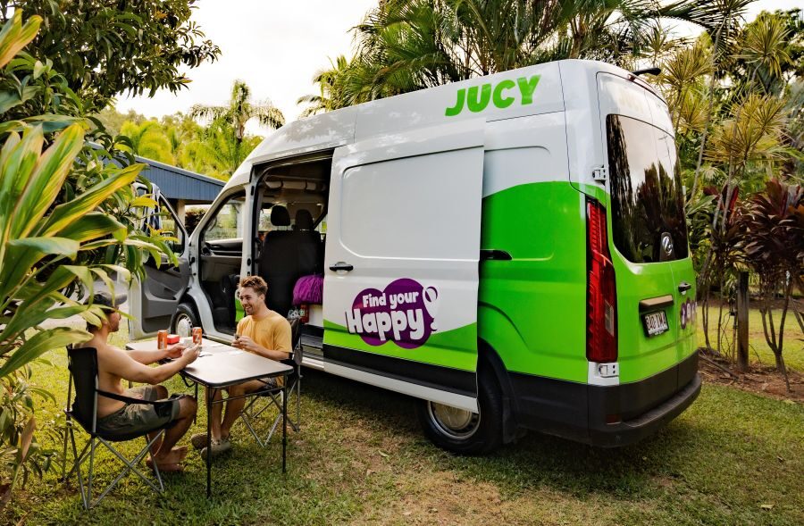 Jucy Coaster staat op een campingplaats met de voor, - en zijdeur open, twee mannen zitten buiten aan tafel