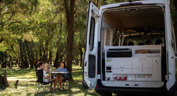 Jucy Coaster geparkeerd tussen bomen op een camping, achterdeuren zijn open en de keuken is te zien, stel zit buiten aan tafel