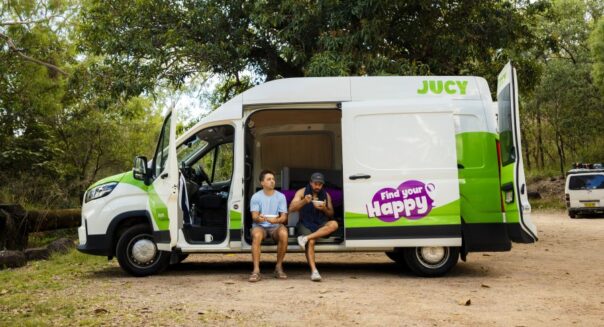 Alle deuren van de geparkeerde Jucy Coaster zijn open, twee mannen eten zittend in de zijdeur