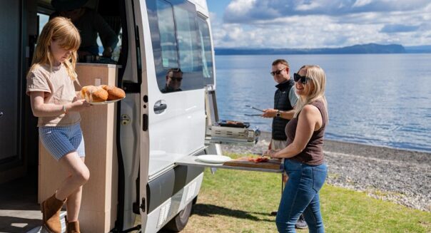 Kind stapt uit camper met bord met broodjes terwijl vader en moeder buiten hamburgers bakken, bij Lake Taupo met Maui Ultima Plus camper