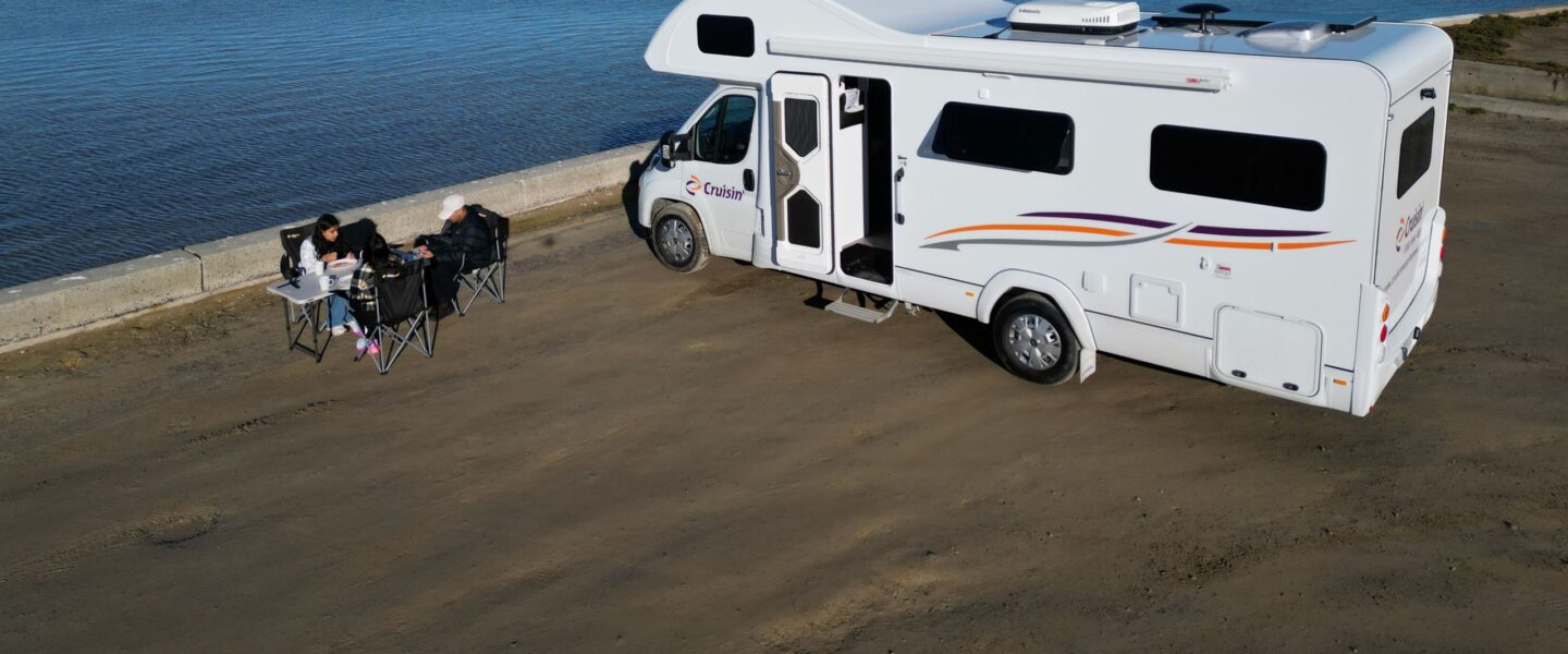 Stel zit in campingstoelen naast Cruisin' 6 Berth Deluxe camper die geparkeerd staat bij het water, in de lucht vliegt een meeuw