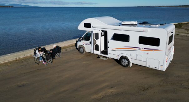 Stel zit in campingstoelen naast Cruisin' 6 Berth Deluxe camper die geparkeerd staat bij het water, in de lucht vliegt een meeuw