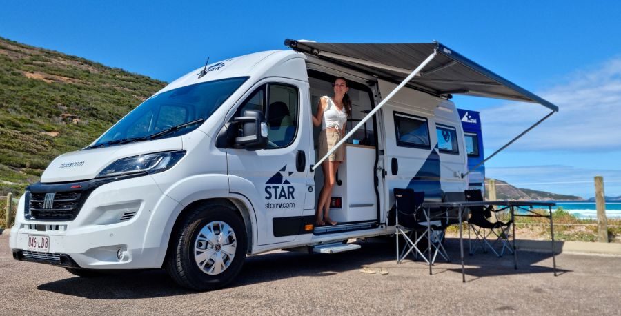 Vrouw staat in de deur van de Star RV Polaris 2, luifel is uitgeklapt en de tafel en stoelen staan buiten