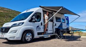 Vrouw staat in de deur van de Star RV Polaris 2, luifel is uitgeklapt en de tafel en stoelen staan buiten