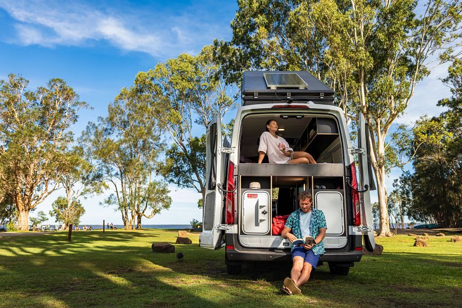 Vrouw ligt op bed en man leest op de bumper van de Britz Voyager camper met de achterdeuren open met