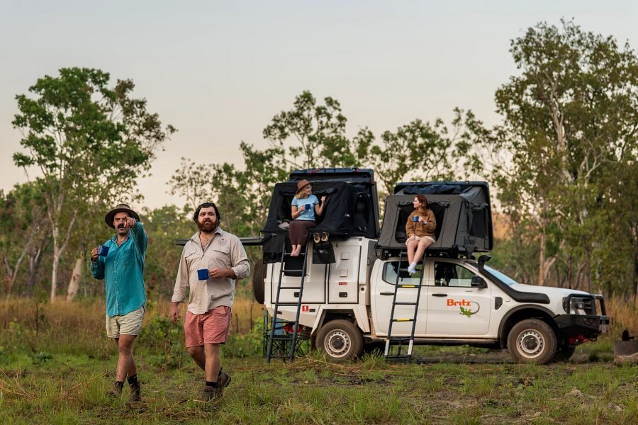 Britz Warrior 4WD Australië rooftop tent uitgeklapt met twee vrouwen zittend in de opening van de tent en twee mannen wandelend met koffie voor de auto