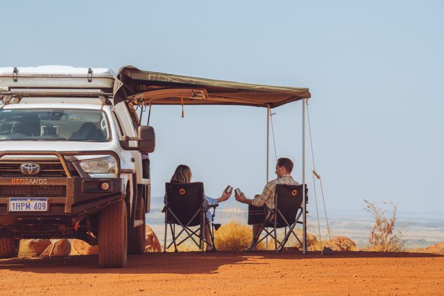 De camperreizigers zitten in hun campingstoelen onder de luifel van hun RedSands 4WD camper en proosten op het fantastische uitzicht