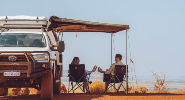 De camperreizigers zitten in hun campingstoelen onder de luifel van hun RedSands 4WD camper en proosten op het fantastische uitzicht