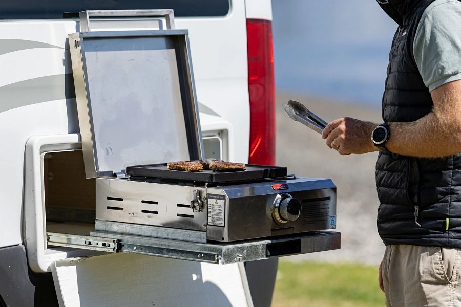 Een man is aan het koken op het uitschuifbare gastoestel aan de buitenkant van de Britz Venturer camper