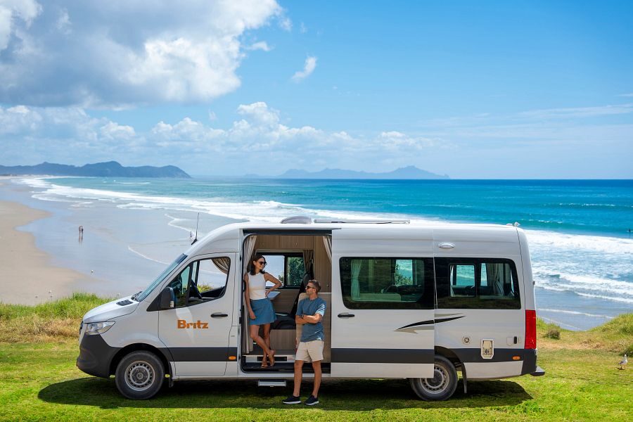 Een koppel staat in de deuropening van de Britz Venturer camper met daarachter uitzicht op de zee in Nieuw-Zeeland