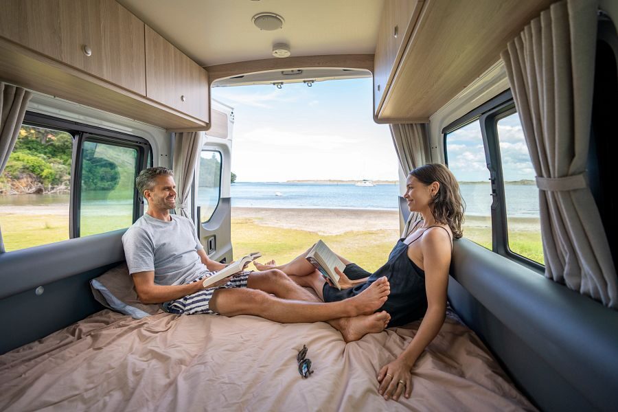Een stel ligt op het tweepersoons bed achterin de Britz Venturer camper in Nieuw-Zeeland een boek te lezen met de achterdeuren open en uitzicht op het water