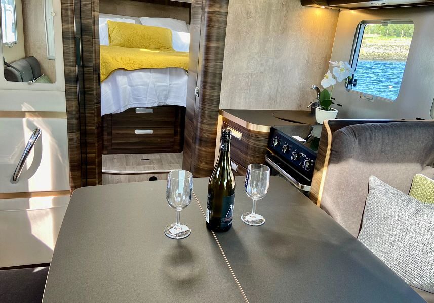 De eettafel met een fleswijn en twee wijnglazen met uitzicht naar de achterkant van de camper waar de slaapkamer is met een vast tweepersoonsbed