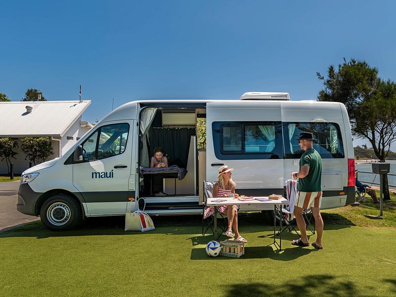 Een koppel zit op de campingstoelen voor de Maui Ultima Plus camper waar een kind binnen op het eenpersoonsbed ligt