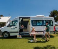 Een koppel zit op de campingstoelen voor de Maui Ultima Plus camper waar een kind binnen op het eenpersoonsbed ligt