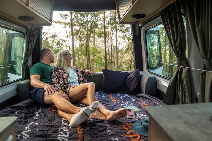 Een koppel zit op het tweepersoonsbed achterin de Maui Ultima Plus camper met de achterdeuren open kijkend naar de natuur buiten