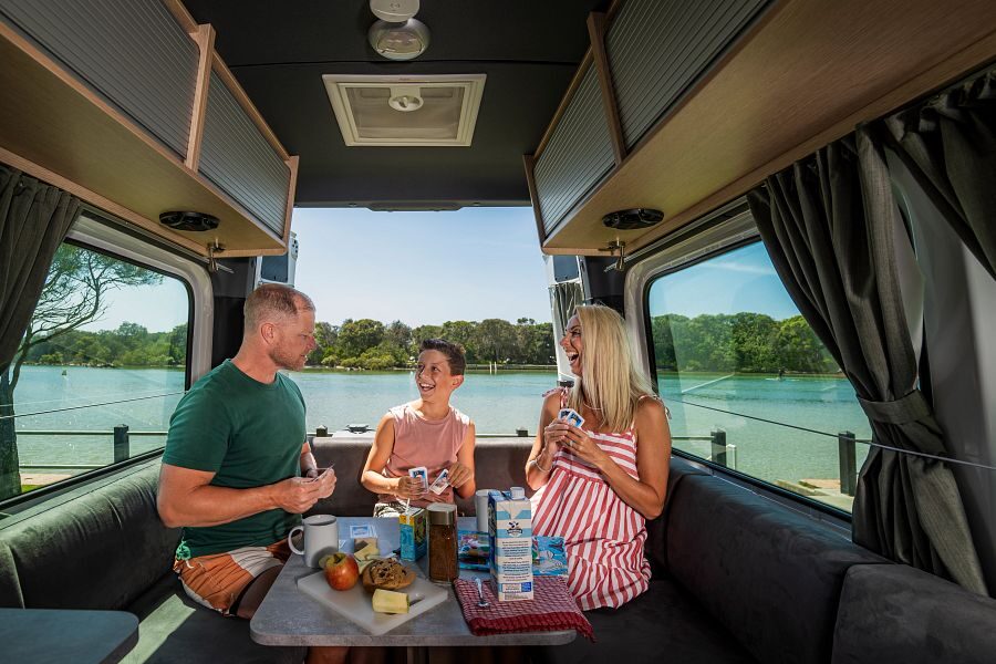Een gezin is aan het ontbijten in de zithoek achterin de Maui Ultima Plus camper met de achterdeuren open en uitzicht op het water