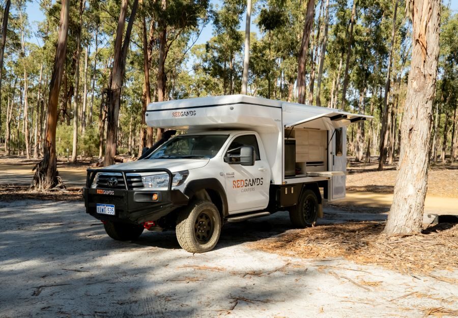 De camper is geparkeerd op een plek tussen de bomen - de klep aan de zijkant is geopend met een uitschuifbaar keukenwerkblad