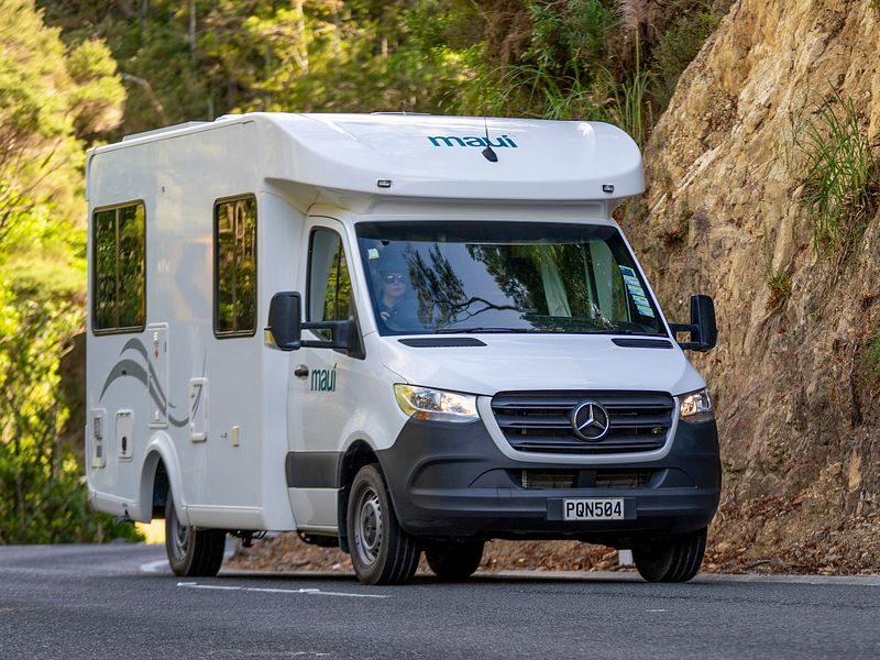 Een vrouw rijdend in de Maui Cascade camper vanaf de voorkant gezien op een weg in Nieuw-Zeeland