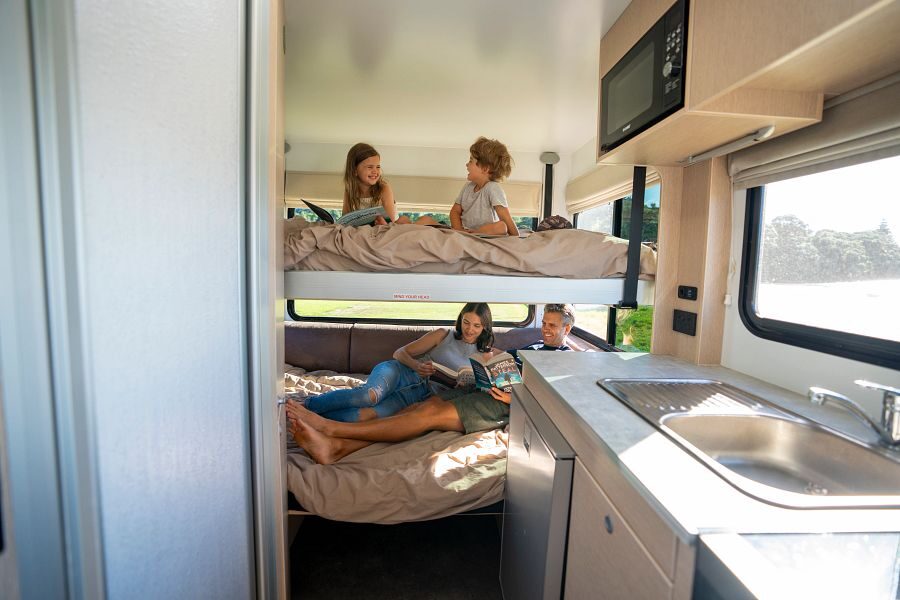 Twee kinderen zitten op het hefbed boven het tweepersoonsbed waar de ouders liggen met een boek achterin de Maui Cascade camper