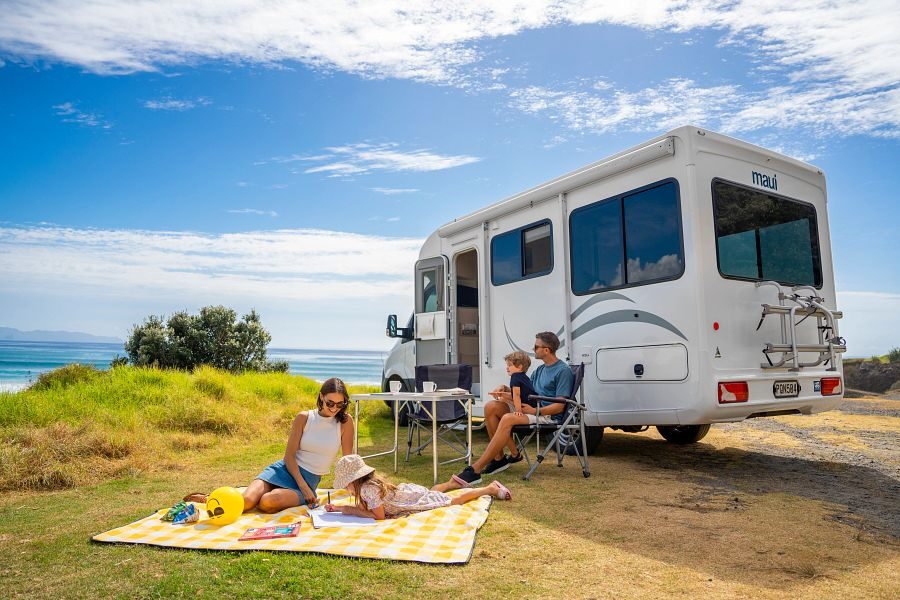 De zijkant en achterkant van de Maui Cascade camper waar een man met een klein kindje op de campingstoel zit en de vrouw met haar dochter op een picknick kleed