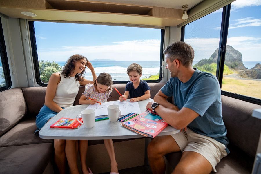 Een gezin met twee kleine kinderen aan het tekenen in de zithoek achterin de Maui Cascade camper met uitzicht op zee in Nieuw-Zeeland