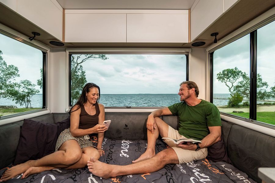 Koppel zit op het tweepersoonsbed achterin de Maui Beach camper met uitzicht op het water