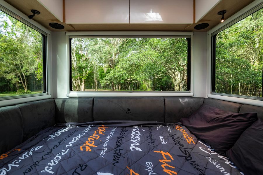Een opgemaakt tweepersoonsbed aan de achterkant van de Maui Beach camper met grote ramen en uitzicht op de natuur