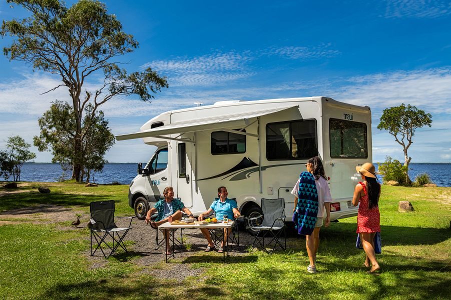 Twee mannen zitten in de campingstoelen onder de luifel van de op het gras geparkeerde Maui Beach camper terwijl er twee vrouwen hun richting op lopen