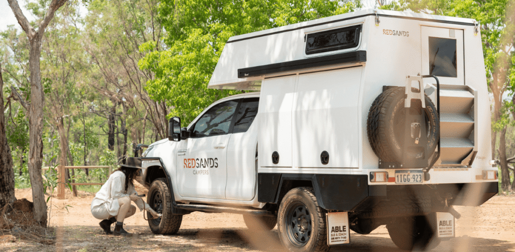De camperreizigster is bezig met het meten van de bandenspanning van de Redsands camper. Aan de achterzijde van de camper is een reservewiel bevestigd en een opklapbaar trapje naar de achterdeur