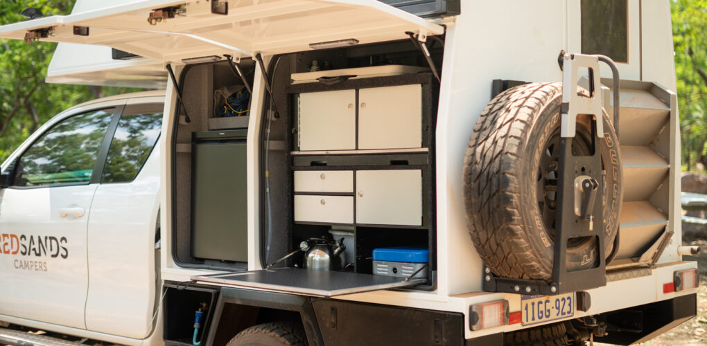 De klep aan de zijkant van de camper is geopend en daarachter bevinden zich onder andere de koelkast, een kooktoestel en opbergladen met kookbenodigdheden en een uitklapbaar werkblad
