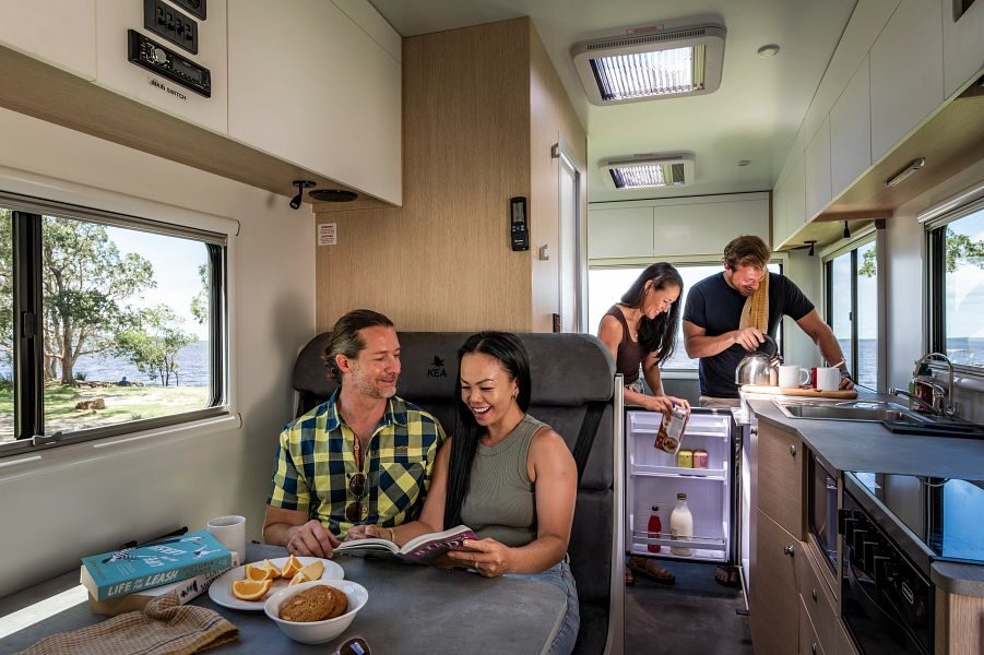 Een koppel zit aan tafel in het middelste zitgedeelte van de Maui River camper terwijl een ander koppel in de keuken staat met de koelkast open om drinken in te schenken