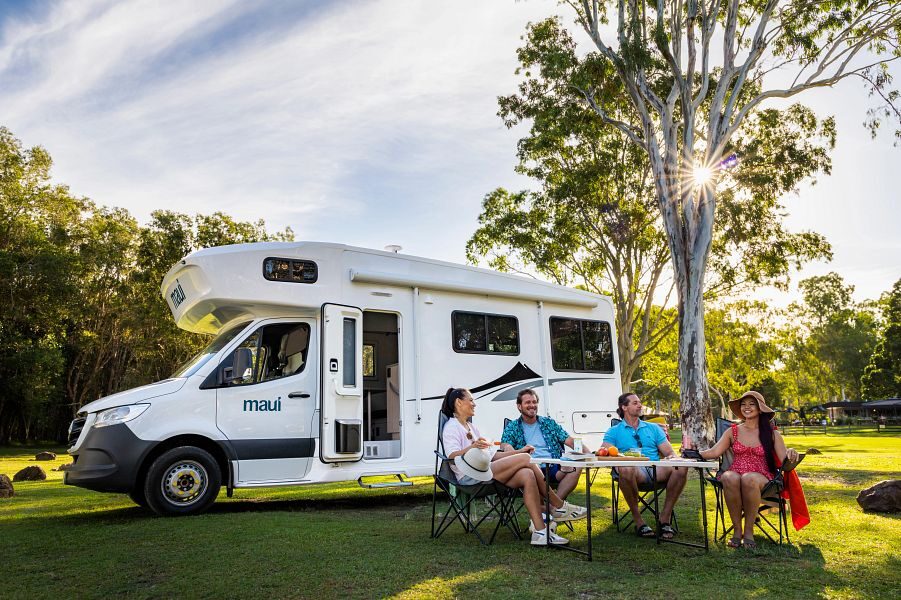 De Maui River camper vanaf de zijkant geparkeerd op het gras naast een boom met voor de camper een vriendengroep zittend op de campingstoelen aan tafel