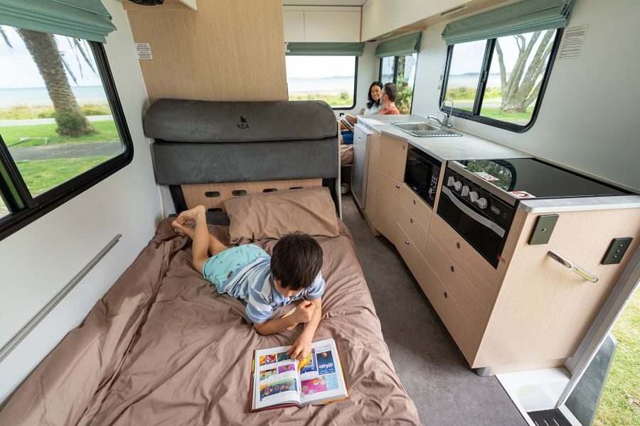 Een kind ligt een boek te lezen op het bed in het midden van de Maui River camper terwijl de ouders achterin de camper aan het praten zijn