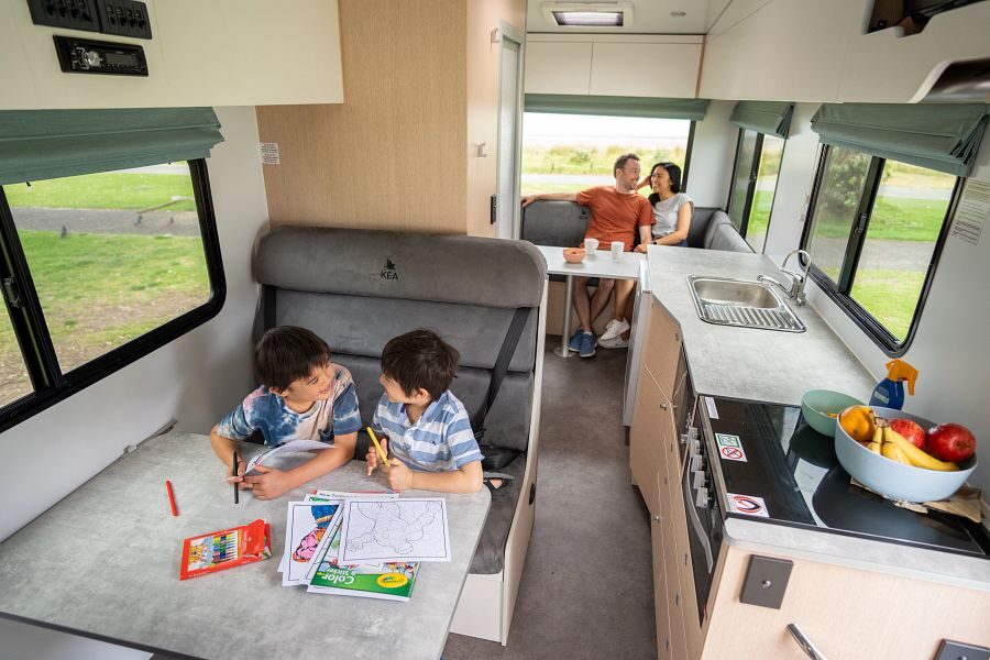 Twee kinderen zijn aan het tekenen aan tafel in het midden van de Maui River camper terwijl de ouders achterin de camper in de zithoek wat aan het drinken zijn