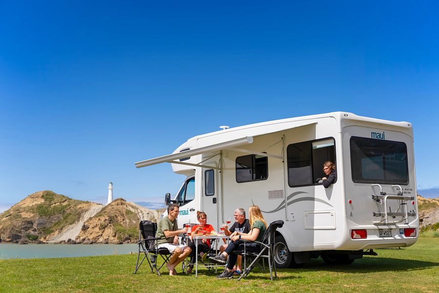 De zijkant en achterkant van de Maui River camper waar een familie onder de luifel zit aan tafel op de campingstoelen en een kind vanuit de zithoek achterin de camper naar buiten kijkt